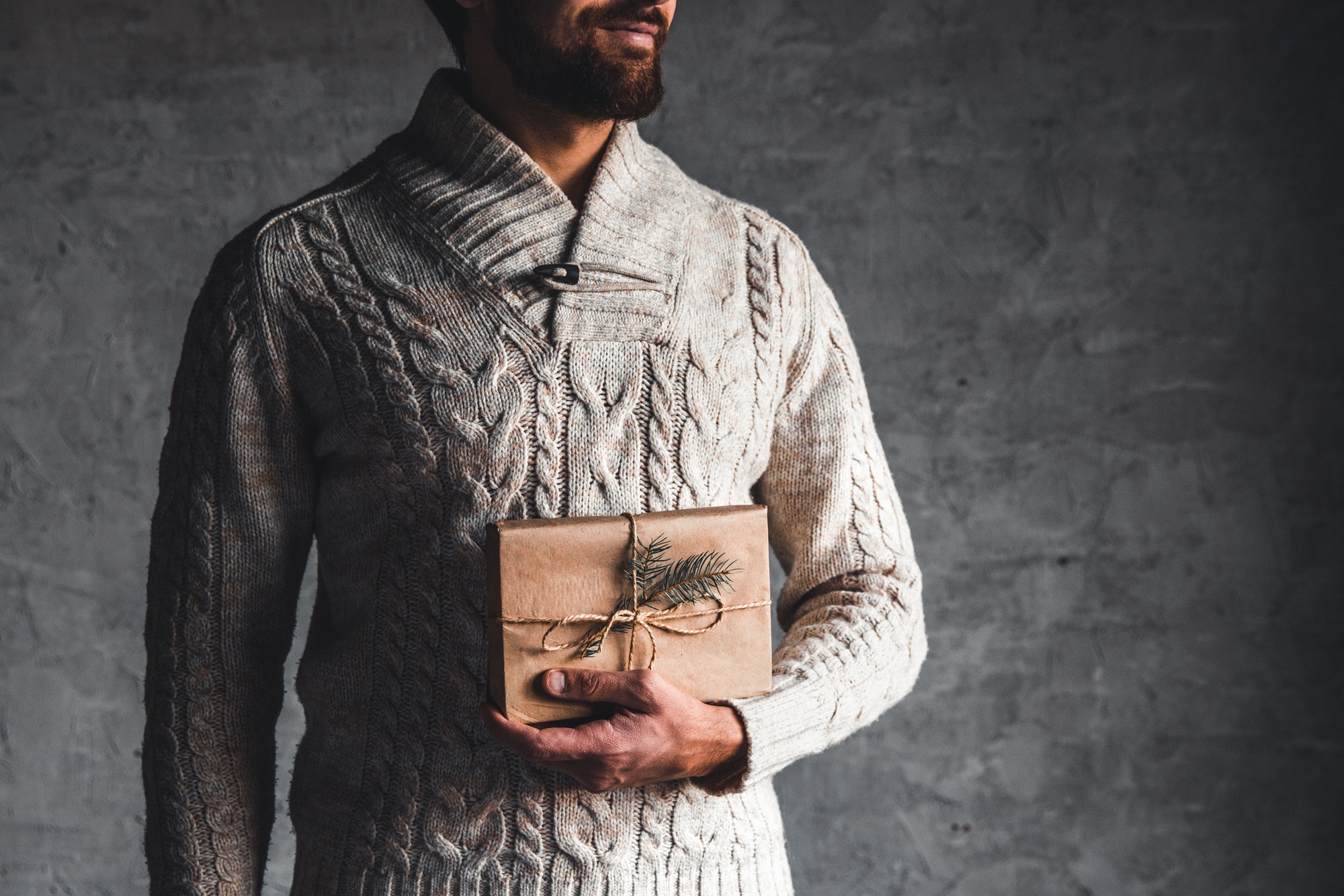 A man holds a craft box for a Christmas present in a beige sweater on a gray background